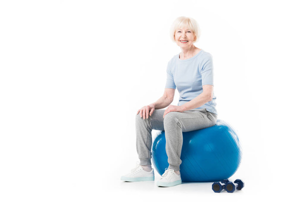 Senior sportswoman sitting on fitness ball with dumbbells on floor isolated on white - Photo, Image