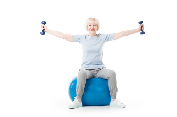 Seniorin mit Kurzhanteln in breiten Händen sitzt auf Fitnessball isoliert auf weißem Untergrund - Foto, Bild