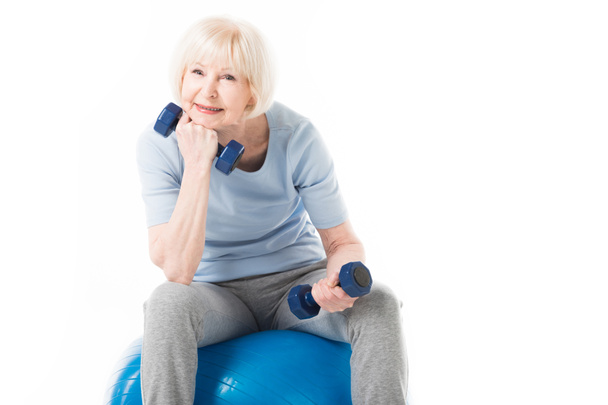 Senior sportswoman sitting on fitness ball with dumbbells in hands isolated on white - Foto, Imagem