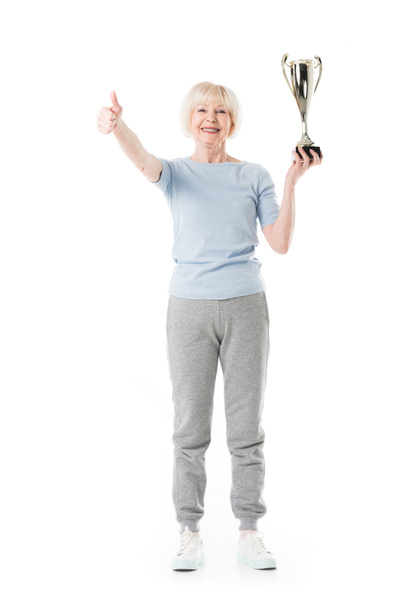 Smiling senior sportswoman showing thumb up gesture and holding trophy isolated on white - Photo, Image