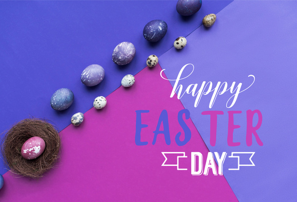 Reihen bemalter Hühner- und Wachteleier und Nest mit fröhlichem Osterfest-Schriftzug auf dem Hintergrund in violetten Tönen - Foto, Bild