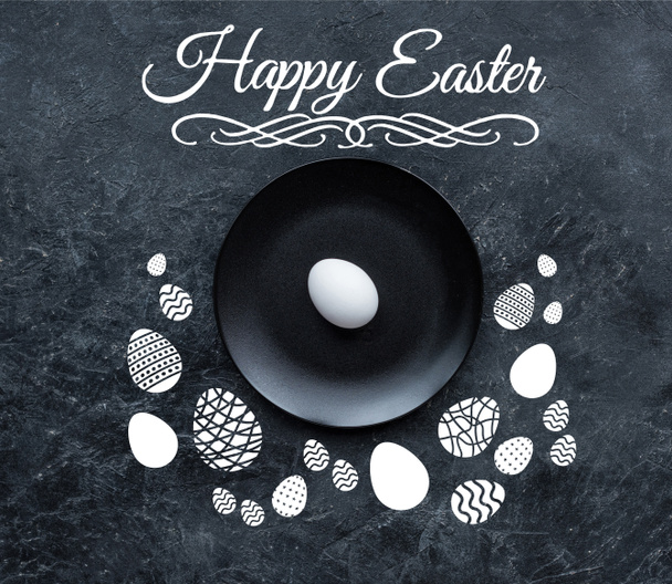White egg on black plate  with happy easter lettering and drawn eggs on dark background - Photo, Image