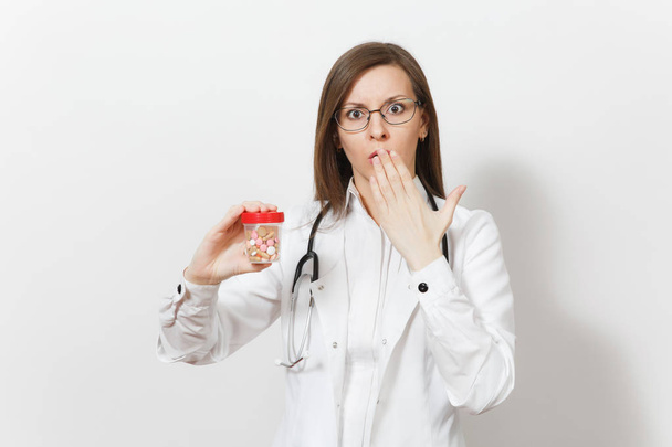 Concerned upset young doctor woman with stethoscope isolated on white background. Female doctor in medical gown holds bottle with pills, put her hand to mouth. Healthcare personnel, medicine concept. - Foto, Imagen