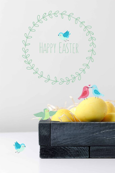 yellow painted easter eggs in wooden box with drawn birds and happy easter lettering on white table - Photo, Image
