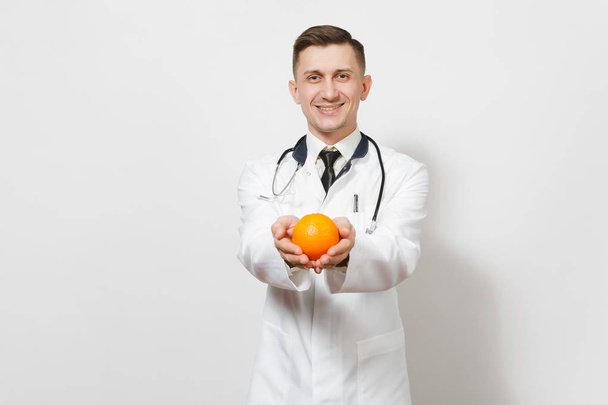 笑みを浮かべてのハンサムな若い医者男は、白い背景で隔離。オレンジを持って医療制服、聴診器の男性医師。医療従事者、健康、医学の概念。適切な栄養。コピー スペース - 写真・画像