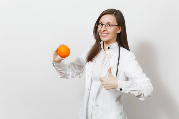 Счастливая красивая молодая женщина врач со стетоскопом, очки изолированы на белом фоне. Женщина-врач в медицинском халате держит оранжевый, показывая большие пальцы вверх. Здравоохранение, медицина
 - Фото, изображение