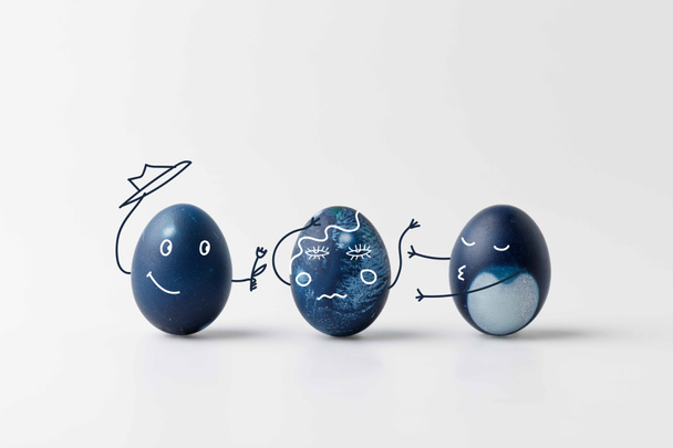 три сині розфарбовані великодні яйця з комічними намальованими обличчями на білій поверхні
 - Фото, зображення