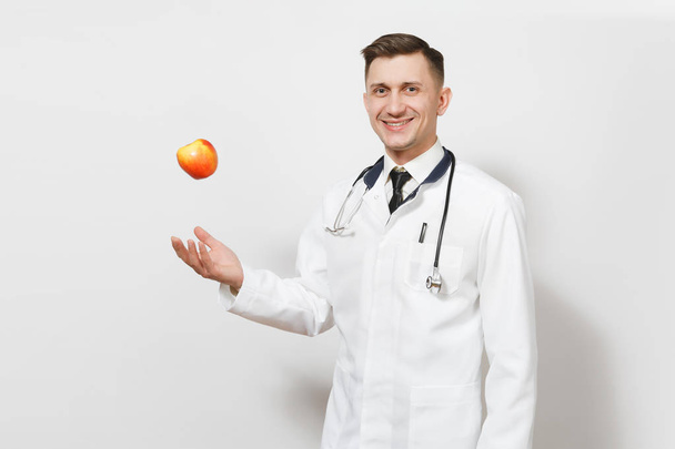 Hymyilevä nuori lääkäri valkoisella pohjalla. Mieslääkäri univormussa, stetoskooppi oksentamassa punaista omenaa. Terveydenhuollon henkilöstö, terveys, lääketiede käsite. Oikea ravitsemus. Kopioi tila
. - Valokuva, kuva