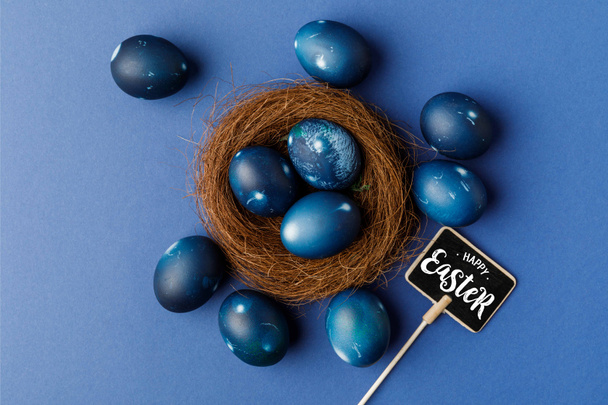 top view of blue painted easter eggs in decorative nest and happy easter lettering on signboard on blue surface - Photo, Image