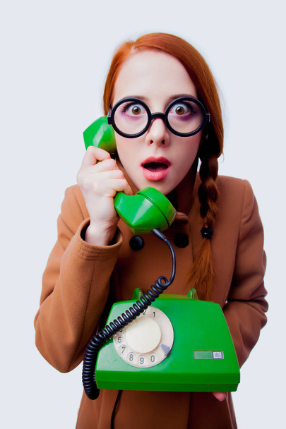 Portrait of a young redhead funny nerd girl with eyeglasses and coat talking by phone on white background - Fotó, kép