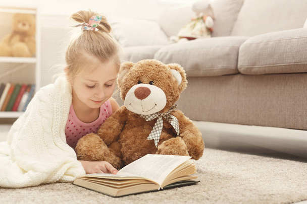 Joyeux petit enfant femelle et son ours en peluche lecture livre sur le sol à la maison
 - Photo, image