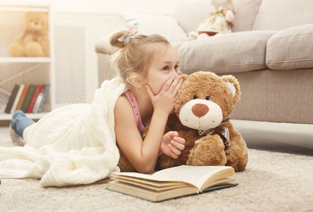 Happy little female child and her teddy bear reading book on the floor at home - Photo, Image