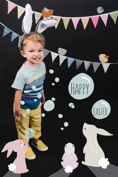 adorable little kid with bunny ears and garland smiling at camera on black, happy easter lettering in bubbles and bunnies collage - Photo, image