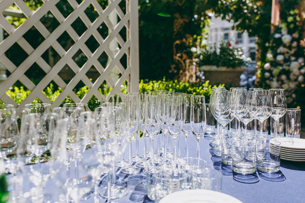 Готовимся к вечеринке. Многие чистые блестящие пустые бокалы для вина, шампанского, виски, сока, воды и других напитков на столе с голубой скатертью в беседке на открытом воздухе партии в саду
 - Фото, изображение