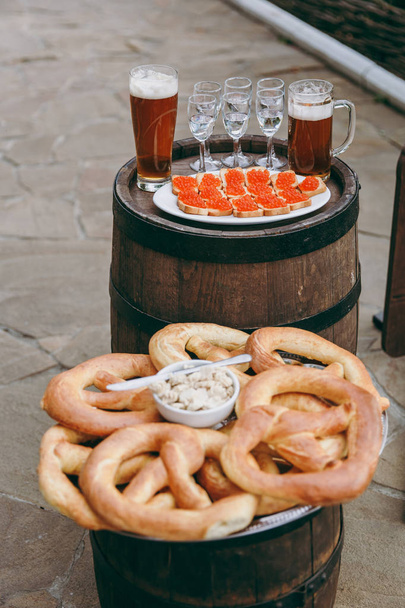 Treats for guests at the oktoberfest. Salted soft baked fragrant pretzels and a spread for them in a plate with a knife, canap with red caviar and glasses with drinks on a tray on a wooden barrels - Photo, Image