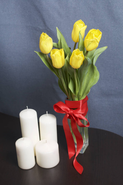 A bouquet of yellow tulips stands in a glass vase wrapped in a red ribbon tied to a bow. Nearby are decorative candles. - Photo, image