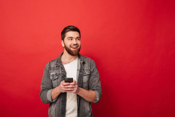 Image of Pleased bearded man holding smartphone and looking away - Foto, Bild