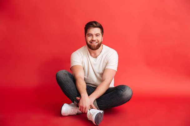 Bearded man sitting on floor isolated over red background - Фото, изображение