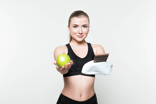Fitness woman holding apple and chocolate bar isolated on white - 写真・画像