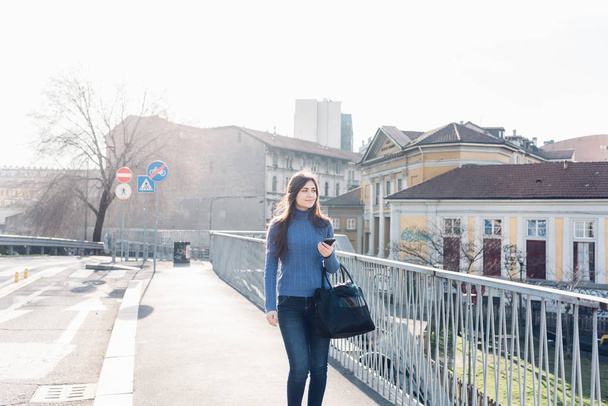 Mujer joven al aire libre caminando en la ciudad luz trasera sosteniendo teléfono inteligente
 - Foto, imagen