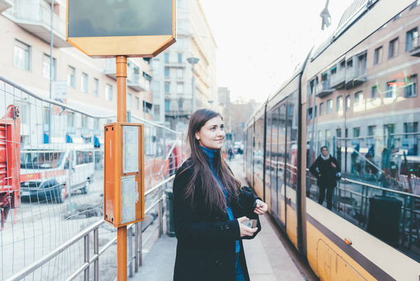 Jovem mulher ao ar livre usando smartphone mão espera paragem de autocarro
 - Foto, Imagem