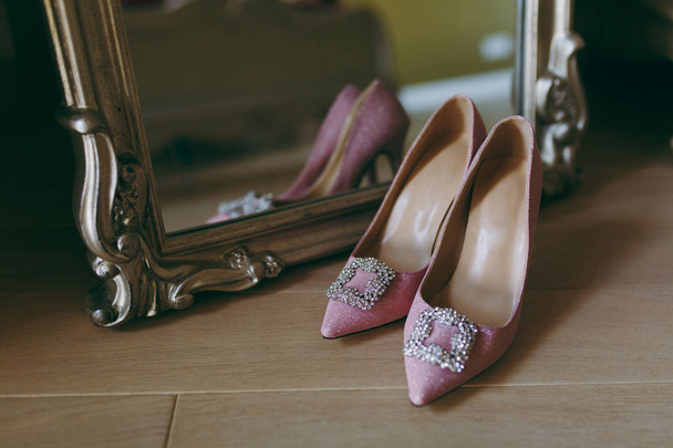 美しいフレームの鏡の近くの木の床の背景に花嫁の鋭い鼻の大きな装飾とエレガントなライト ピンク結婚式繊維の光沢のある靴を閉じます。結婚式の摩耗、アクセサリー - 写真・画像