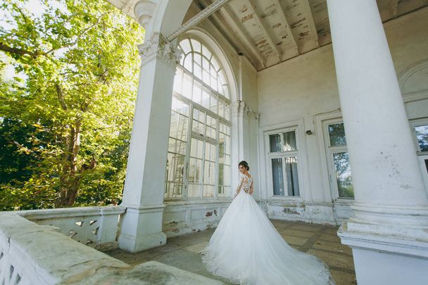 Hermosa sesión de fotos de boda. Joven novia en un vestido de encaje blanco con una pluma larga con un exquisito peinado en el interior vintage en la terraza de una casa vieja con columnas cerca del jardín
 - Foto, Imagen