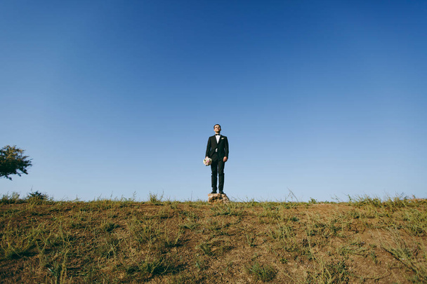 Beautiful wedding photosession. Handsome bridegroom in a black suit and white shirt with a bouquet of the bride stands on a stone on walk around the big green field against blue sky background - 写真・画像