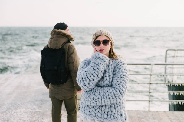 attractive stylish girl in sunglasses and merino wool sweater on winter quay, boyfriend standing behind and looking at sea - Foto, immagini