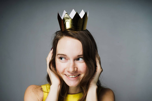 happy young girl in a yellow dress closed her ears, smiling, on her head a golden crown - Photo, Image