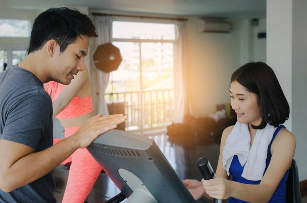 asian young woman slim body exercising on bicycle machine with young man personal trainer in fitness gym, bodybuilder, healthy lifestyle, fitness, workout and sport training concept, soft focus - Photo, Image