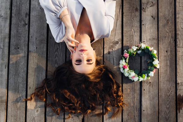 Eine Frau auf einer Holzfläche mit geschlossenen Augen genießt eine Ruhepause. ein Blumenkranz liegt neben einer schönen Nymphe - Foto, Bild