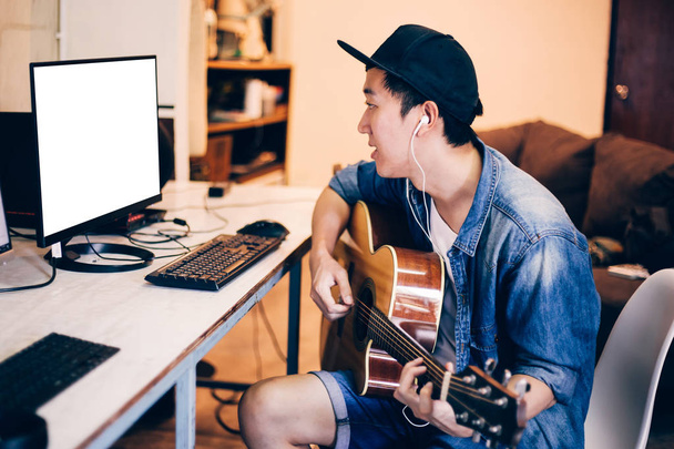 Young Asian man learning how to play guitar on computer monitor. Male guitarist watching online tutorial. Include clipping path on monitor - Photo, Image