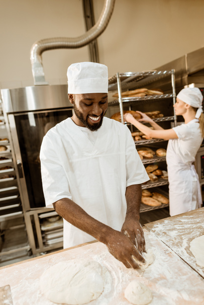 african american baker kneading dough at baking manufacture while his colleague working blurred on background - Foto, Imagen