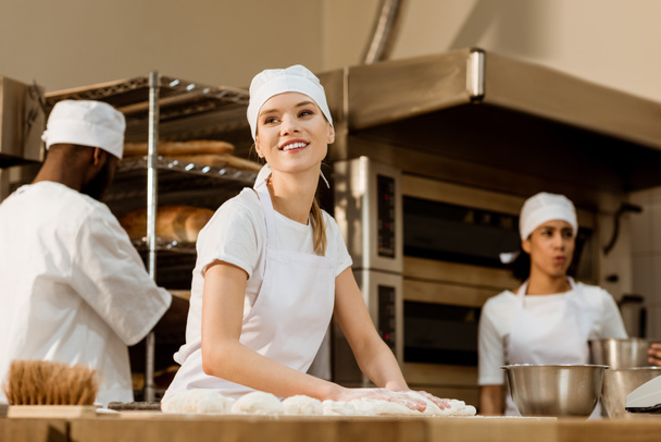 молода жінка-пекарня замішує тісто на виробництві випічки, в той час як її колеги працюють розмиті на фоні
 - Фото, зображення