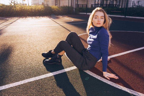 beautiful stylish blond girl in a blue sweater sits on the sports field posing - Foto, afbeelding