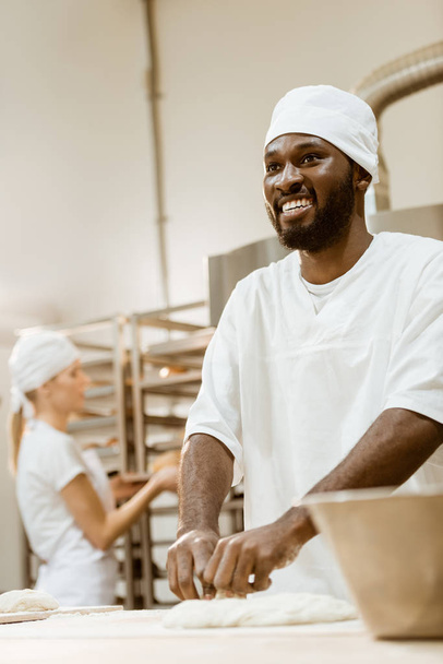 happy african american baker kneading dough while his female colleague working on background at baking manufacture - Fotografie, Obrázek