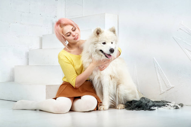 attractive young girl with short blond hair in a yellow sweater and skirt hugs her beloved pet - a dog breed Samoyed - Photo, image