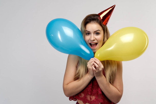 A young cheerful long-haired girl with a cap on her head and with balloons in her hands smiles happily opening her mouth.  - Foto, Imagem