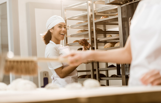 junge Bäckerin im Gespräch mit Kollegin in Backmanufaktur, während sie Arbeitsplatz von Mehl reinigt - Foto, Bild