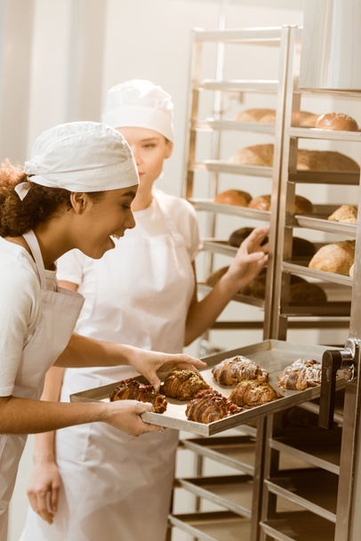 panaderos femeninos jóvenes poniendo pastelería fresca en los estantes en la fabricación de hornear
 - Foto, Imagen