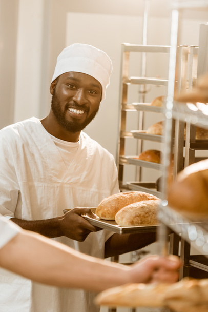 улыбающийся африканский пекарь с хлебом на подносе при выпечке
 - Фото, изображение