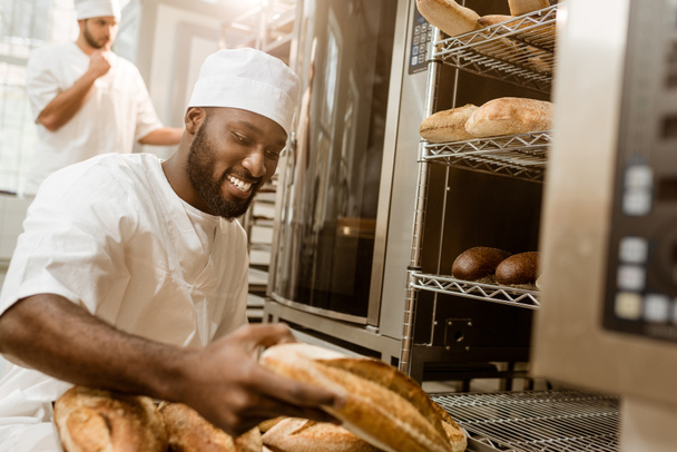 happy african american baker with tray of fresh loaves of bread on baking manufacture - Photo, Image