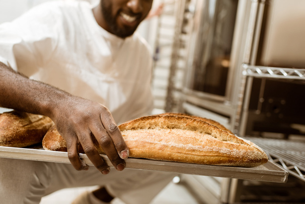 bijgesneden schot van Afro-Amerikaanse bakker met lade van verse broden op bakken vervaardiging - Foto, afbeelding