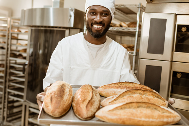 guapo panadero afroamericano con bandeja de panes frescos en la fabricación de hornear
 - Foto, Imagen