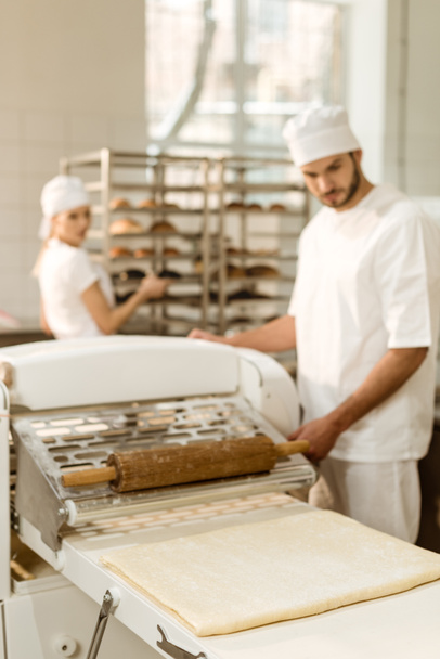 apuesto panadero joven que trabaja con rodillo de masa industrial en la fabricación de la hornada
 - Foto, Imagen