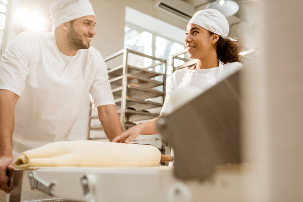 panaderos jóvenes felices trabajando con rodillo de masa industrial en la fabricación de hornear
 - Foto, Imagen