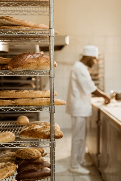 afro-amerikai baker munkahelyen, polcok, friss kenyér, előtérben a nyers tészta elkészítése - Fotó, kép