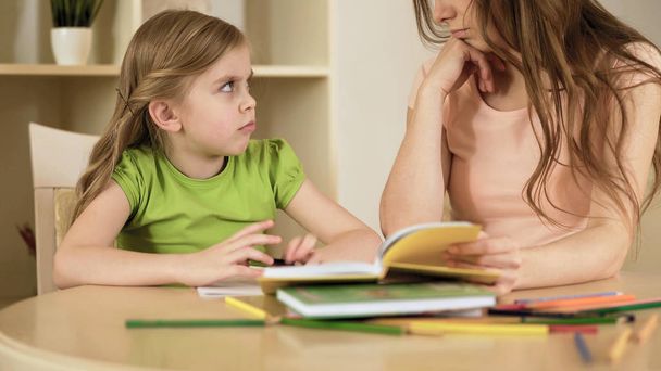 Upset little girl with misunderstanding looking at her mother doing school tasks - 写真・画像