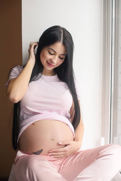 Portrait of smiling pregnant woman sit in window sill - Photo, Image